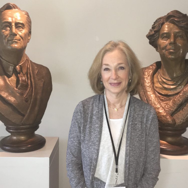 Judy Winzemer, a white woman wearing a grey sweater, standing in front of busts of Franklin and Eleanor Roosevelt