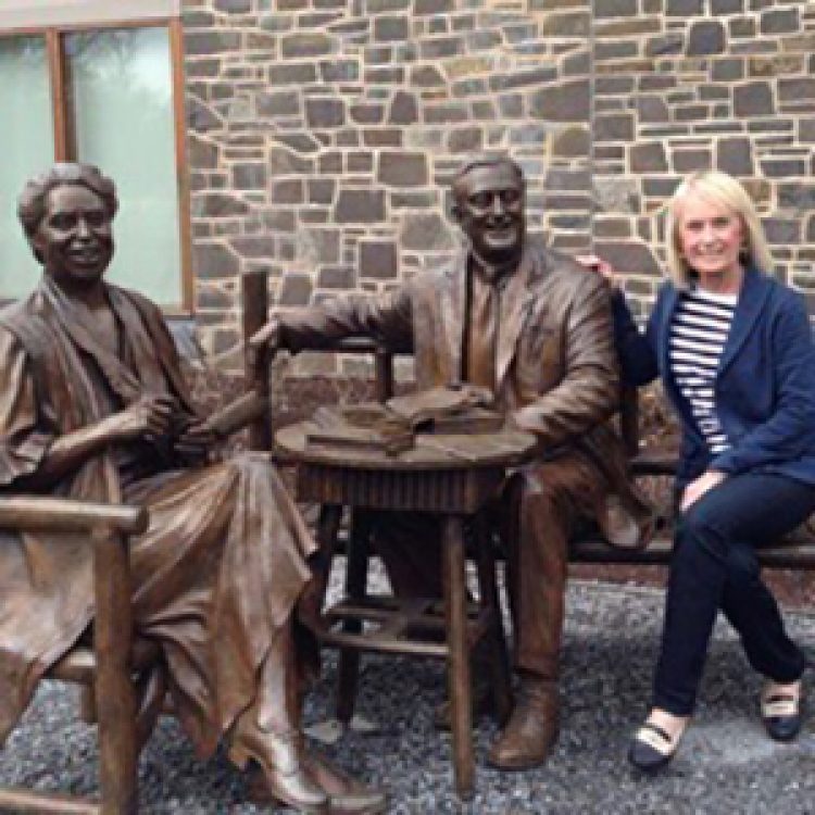Nancy Chando, a white woman with blonde hair, sits on a bench with Franklin and Eleanor Roosevelt statues at the FDR Presidential Library