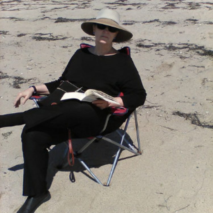 Kathy Roe, wearing all black and a wide brim hat sitting on the beach and reading a book,