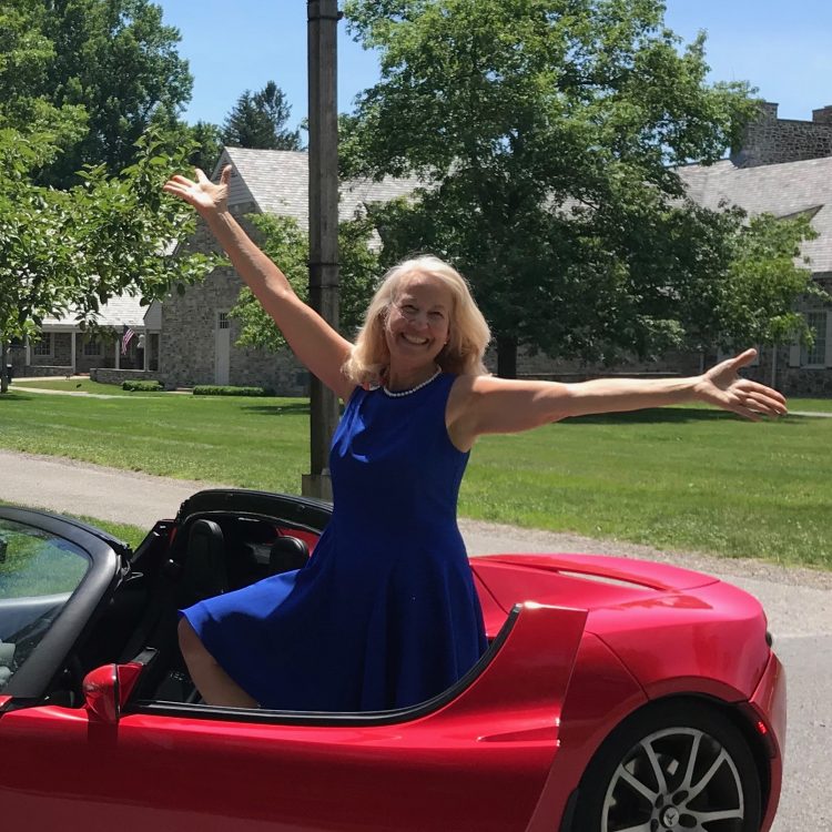 Image of Anne Sumers, a white woman with blond hair, wearing a blue dress and sitting in a convertible car with her arms outstretched.