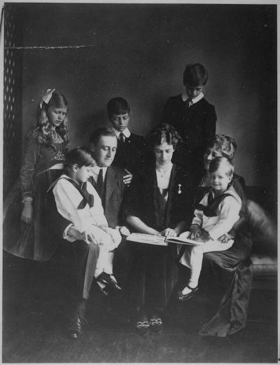 Black and white photo of Franklin and Eleanor Roosevelt with Sara Delano Roosevelt and their children, Franklin Jr., Anna, Elliott, James, and John gathered around a book.