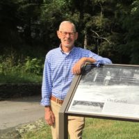 Roger Kolp, a white man wearing a plaid button up, stands next to a sign at Hyde Park
