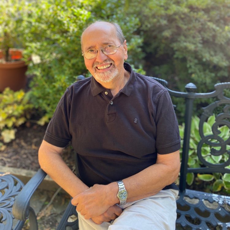 Picture of Sal Assenza, a white man wearing a black polo shirt, sitting on a bench in a garden
