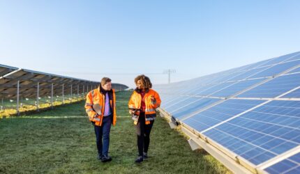 Male and female engineers working at solar power plant. Two technicians in reflective clothing walking between rows of photovoltaic panels at solar farm.