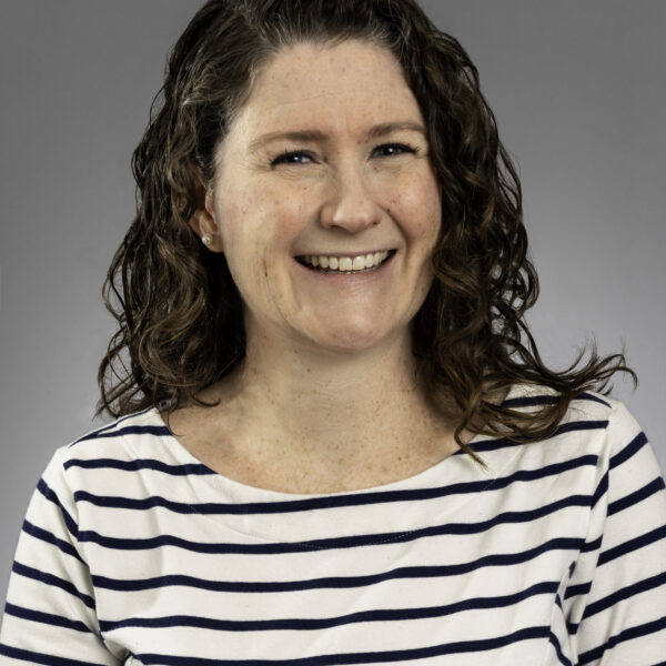 Woman with curly brown hair, wearing a white and navy striped shirt, smiles at the camera against a plain gray background.
