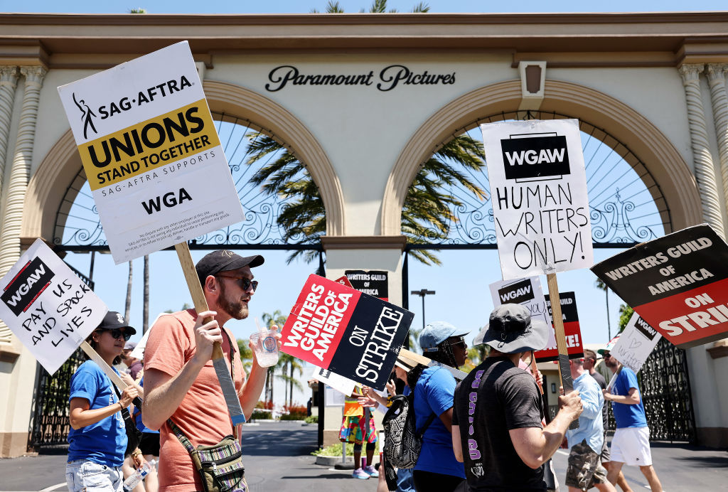 People picket outside the Paramount Pictures gate holding signs supporting the Writers Guild of America strike, with messages like “WRITERS GUILD ON STRIKE” and “HUMAN WRITERS ONLY!” under a sunny sky.