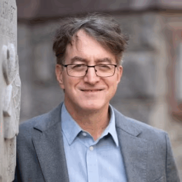 Professor Victor Pickard. A middle-aged man with glasses and tousled brown hair, wearing a light blue shirt and grey blazer, is smiling outdoors in front of a stone wall.
