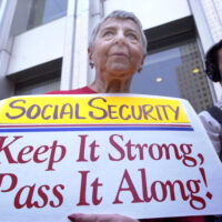 An older woman holds a sign reading Social Security: Keep It Strong, Pass It Along! during a public demonstration, with several people and tall buildings visible in the background.