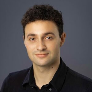 A young man with short, curly dark hair and light skin wears a black collared shirt and smiles softly against a plain gray background.