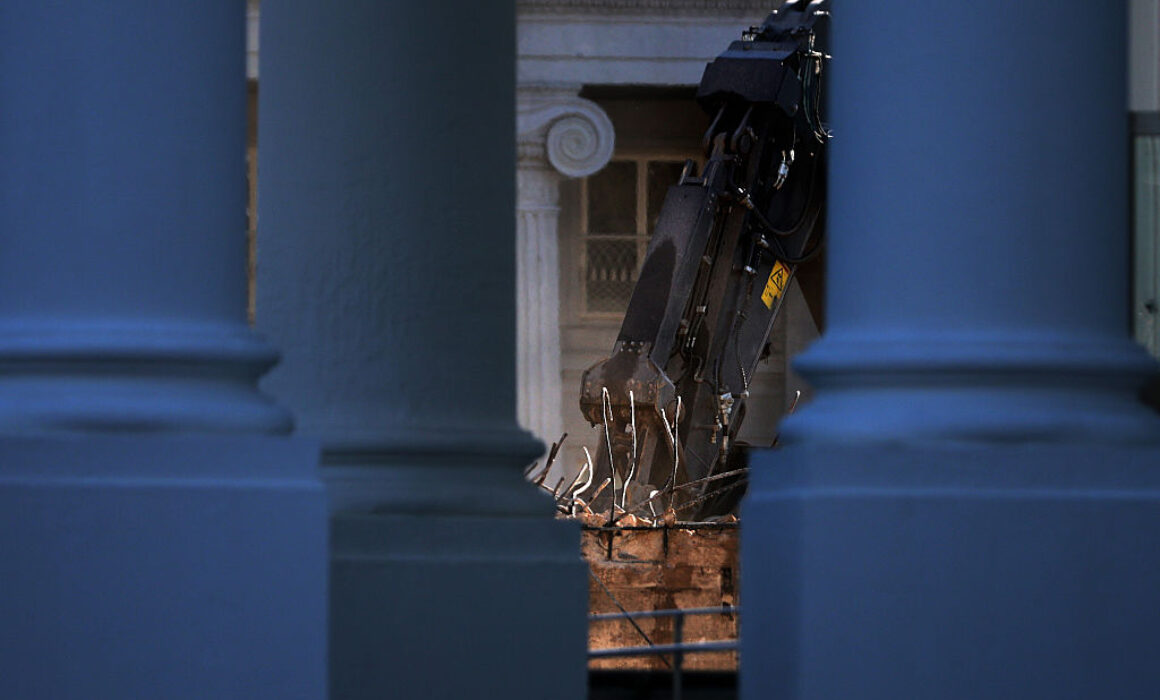 A construction excavator with a claw attachment is seen through the gaps between large blue columns, with a classical-style building featuring columns and a decorative frieze in the background.