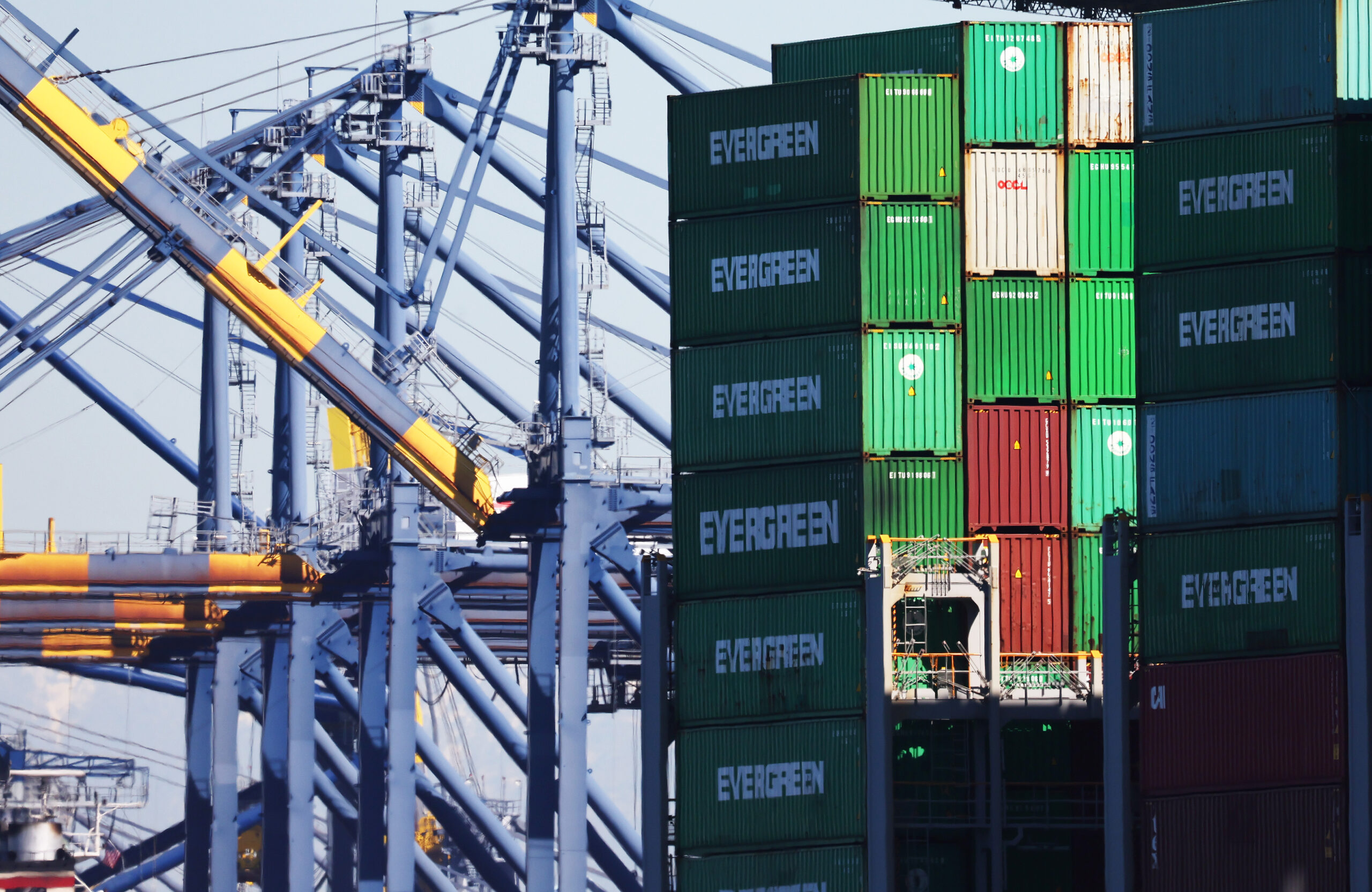 Tall stacks of green shipping containers labeled EVERGREEN stand next to blue and yellow cranes at a busy shipping port under a clear sky.