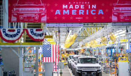 A row of white trucks on an assembly line in a factory. Above them is a large red banner reading A FUTURE MADE IN AMERICA with the White House logo. American flags and patriotic bunting decorate the area.