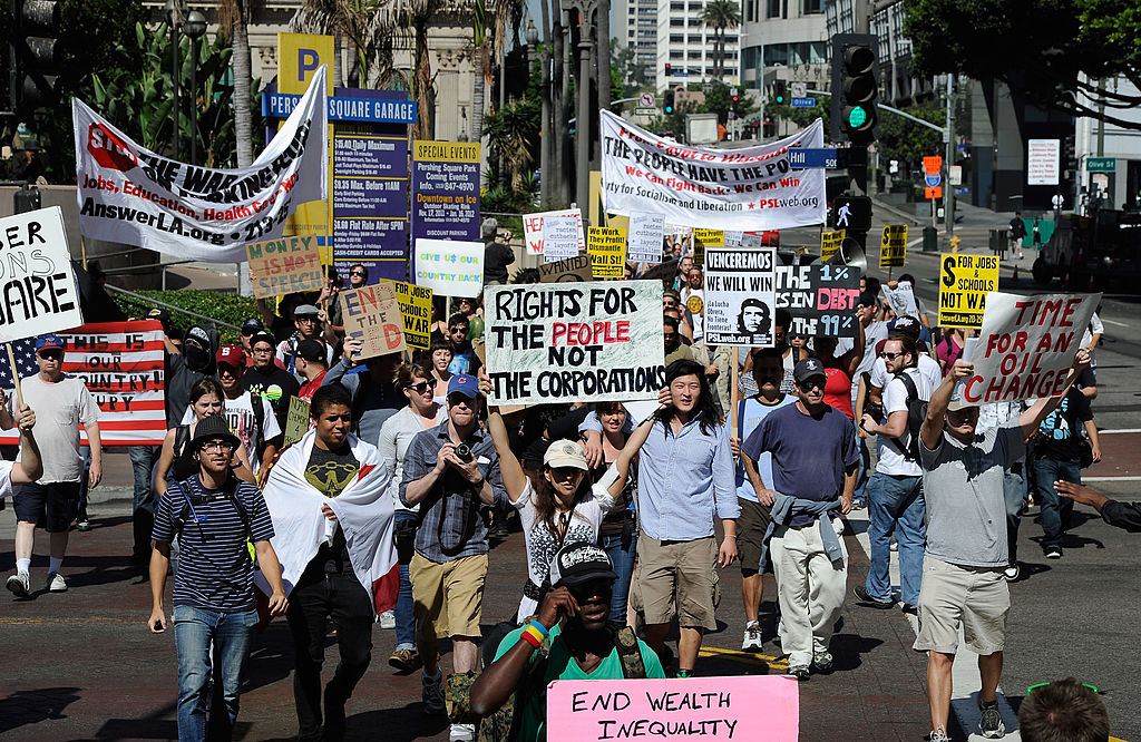 A diverse group of protesters march in a city holding signs with messages like “Rights for the People Not the Corporation” and “End Wealth Inequality.” Banners and buildings are visible in the background.