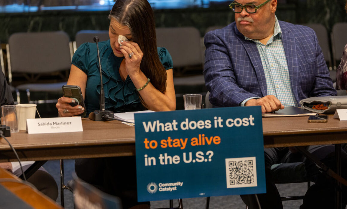 A woman wipes her eyes while looking at her phone during a meeting. Next to her sits a man in glasses. A sign on the table reads, What does it cost to stay alive in the U.S.? with a QR code and Community Catalyst branding.