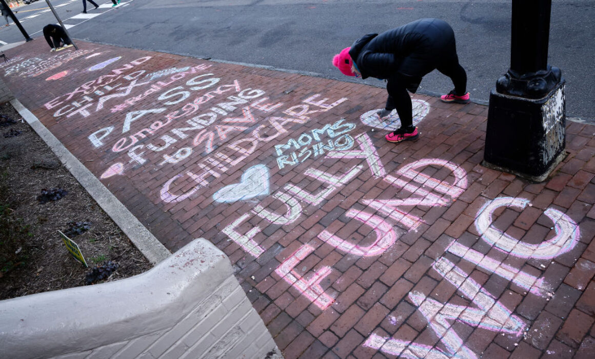 A person writes colorful chalk messages on a brick sidewalk, including phrases like Pass emergency funding to save childcare and Fully fund WIC. Another person writes further down the sidewalk.