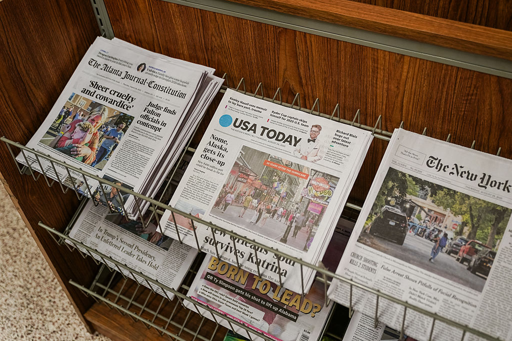 Newspapers, including The Atlanta Journal-Constitution, USA Today, and The New York Times, are displayed on metal racks in a wooden shelving unit. Headlines and photos are visible on the front pages.