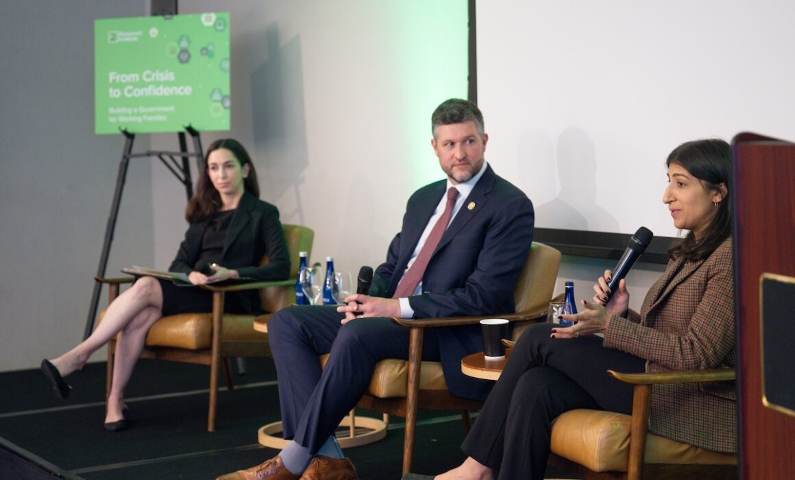 Three people in business attire sit on stage during a panel discussion. A woman on the right speaks into a microphone, while the other two listen. A sign in the background reads “From Crisis to Confidence.”.