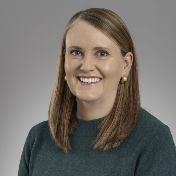 A woman with straight, shoulder-length light brown hair and gold earrings smiles at the camera. She is wearing a green, long-sleeve top and is posed in front of a plain, light gray background.