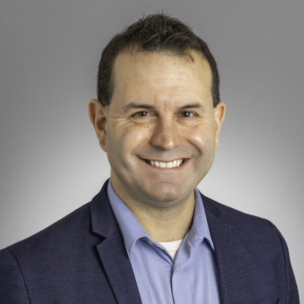 A man with short dark hair, wearing a blue blazer over a light blue collared shirt, smiles at the camera against a plain gray background.