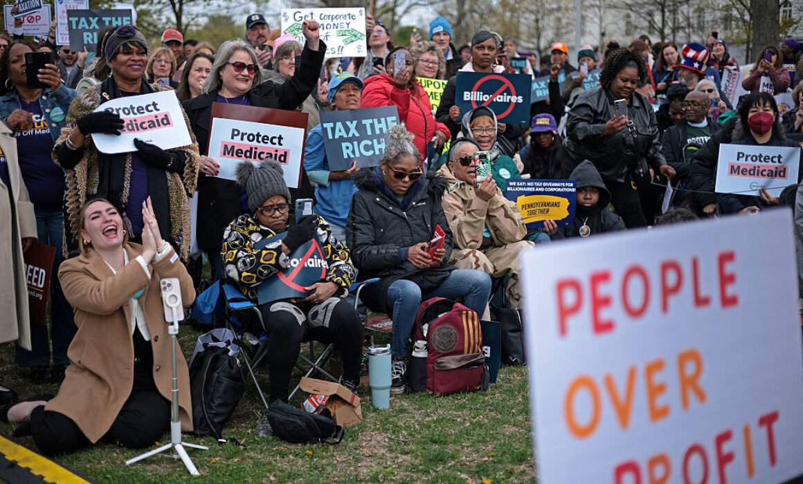 A diverse group of people at an outdoor rally hold signs reading Protect Medicaid and Tax The Rich. A large sign in the foreground says People Over Profit. The crowd appears engaged and supportive.