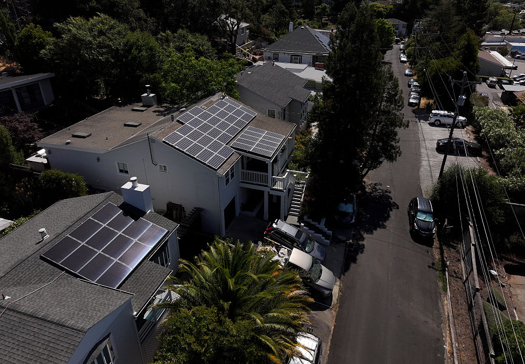 Aerial view of a residential neighborhood with houses featuring solar panels on their rooftops, surrounded by trees, parked cars, and a quiet street on a sunny day.