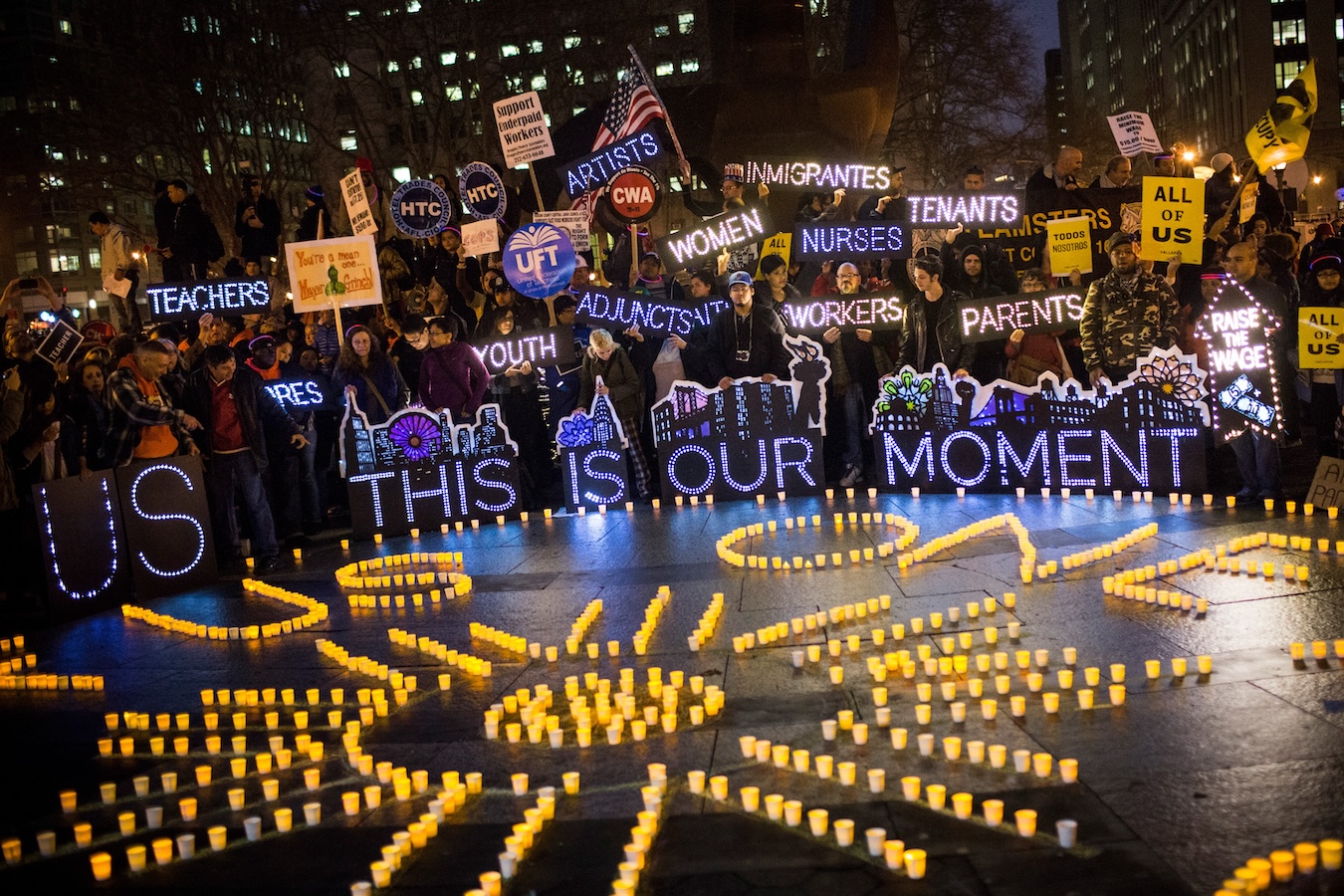 A nighttime protest features people holding illuminated signs and placards supporting teachers, artists, immigrants, nurses, youth, tenants, and more. Candles on the ground form sunbeams and the words US and WE ARE ONE.