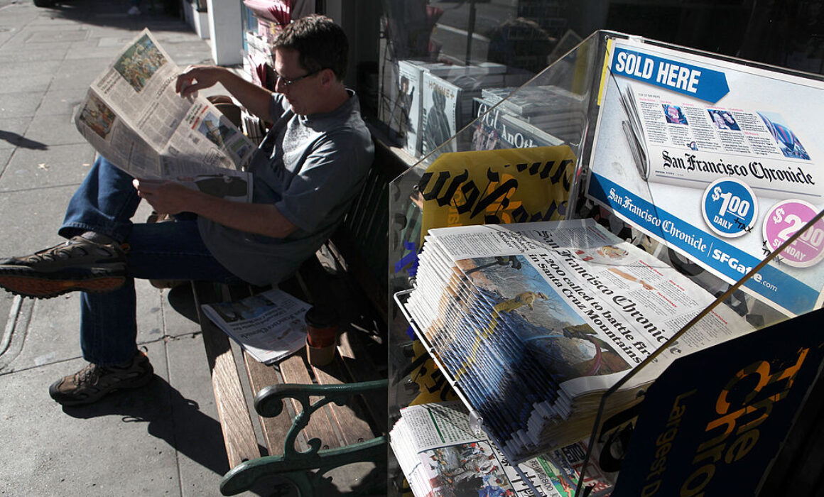 A man sits on a bench outside, reading a newspaper. Nearby, a display rack holds copies of the San Francisco Chronicle for sale, with signs advertising the publication and its price.