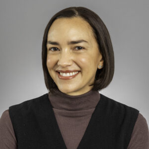 A woman with straight dark brown hair in a bob, wearing a black sleeveless top over a mauve turtleneck, smiles in front of a plain gray background.