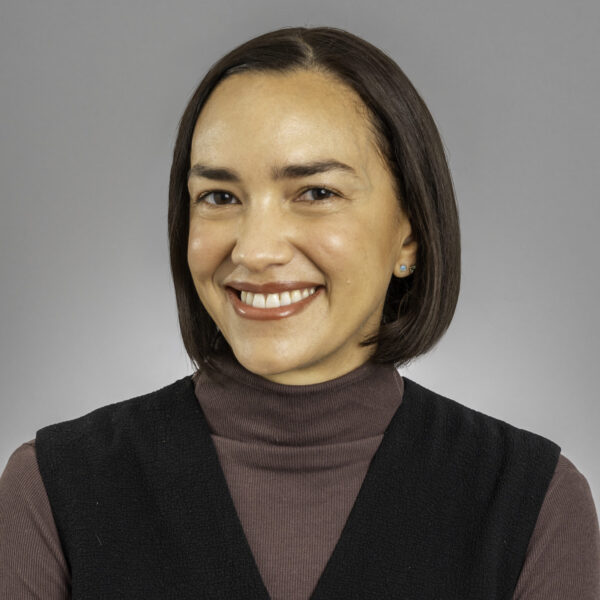 A woman with straight dark brown hair in a bob, wearing a black sleeveless top over a mauve turtleneck, smiles in front of a plain gray background.