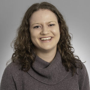 A woman with curly brown hair and fair skin smiles at the camera. She is wearing a gray, textured, cowl-neck sweater and is posed against a plain gray background.