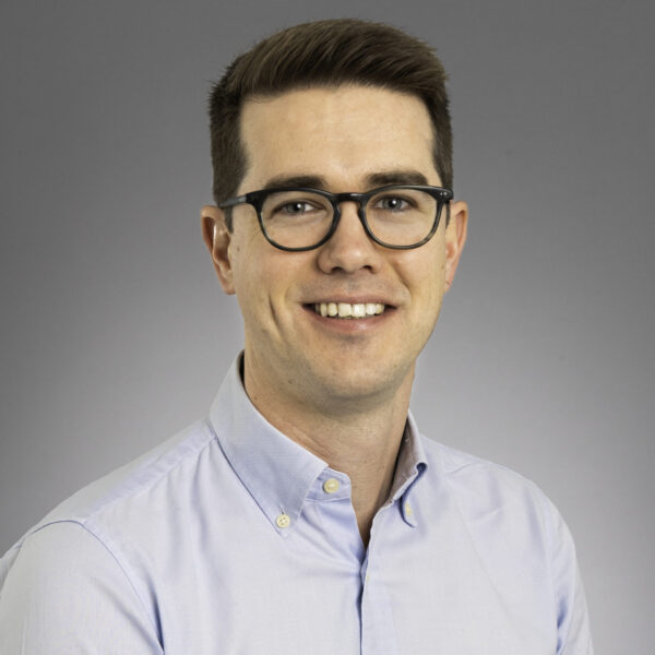 A man with short brown hair and glasses, wearing a light blue button-up shirt, smiles at the camera against a plain gray background.