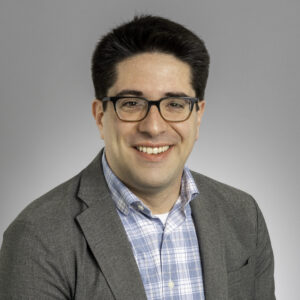 A man with short dark hair and glasses, wearing a gray blazer over a blue and white plaid shirt, smiles at the camera against a plain light gray background.
