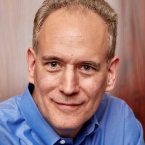 A middle-aged man with short, light brown hair and blue eyes, wearing a blue button-up shirt, smiles gently at the camera. The background is blurred with reddish-brown vertical lines.