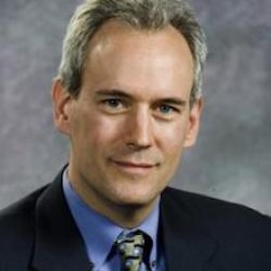A middle-aged man with short gray hair, wearing a dark suit jacket, blue dress shirt, and patterned tie, poses in front of a gray, softly blurred background.