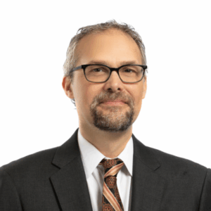 A man with short, graying hair, a beard, and glasses is wearing a dark suit, white shirt, and a patterned tie, standing against a plain white background.