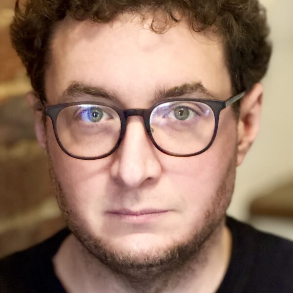 A man with short curly brown hair, wearing black glasses and a black shirt, looks directly at the camera with a neutral expression. A blurred brick wall is in the background.