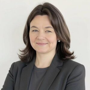 A woman with shoulder-length brown hair, wearing a dark blazer and gray top, stands against a plain light background with her arms crossed, smiling confidently.