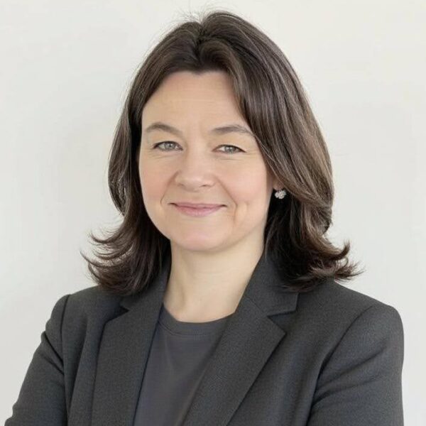 A woman with shoulder-length brown hair, wearing a dark blazer and gray top, stands against a plain light background with her arms crossed, smiling confidently.