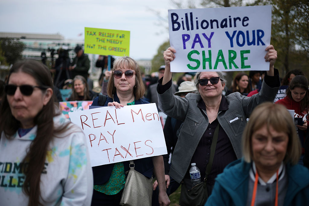 A group of people at a protest hold signs reading REAL Men PAY TAXES and Billionaire PAY YOUR SHARE. Other people and protest signs are visible in the background.