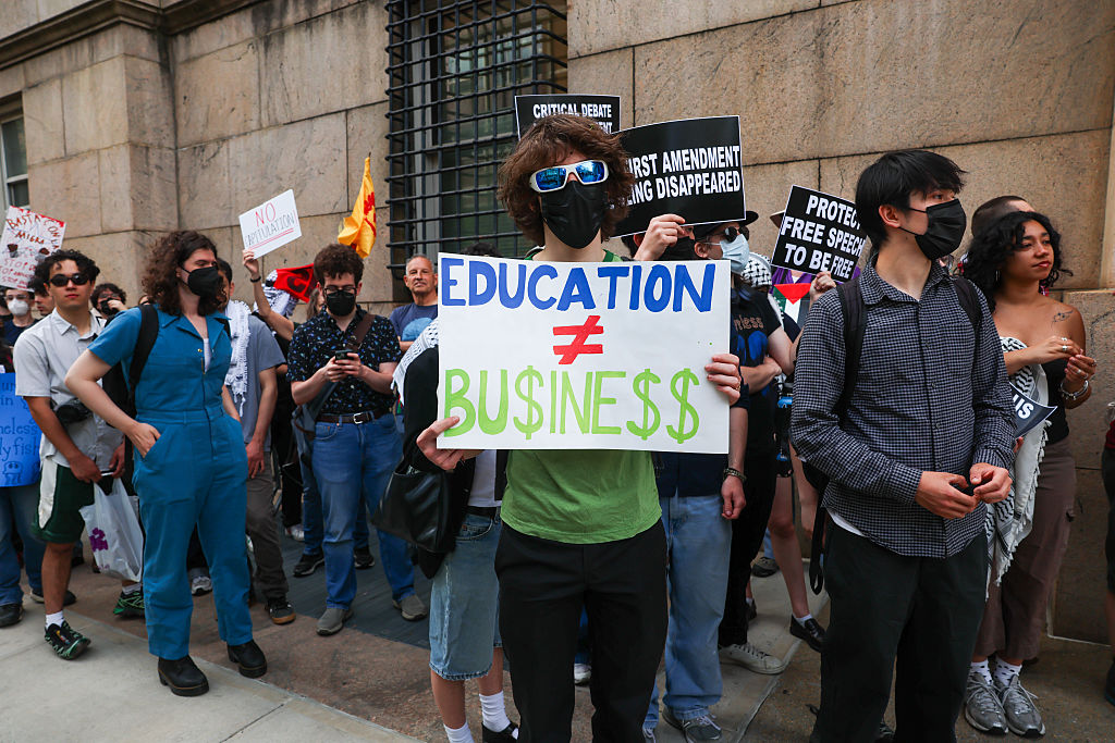 A group of people protest outside a building. One person stands at the center holding a sign that reads EDUCATION ≠ BU$INE$$ in blue and green letters. Several protesters wear masks and sunglasses.