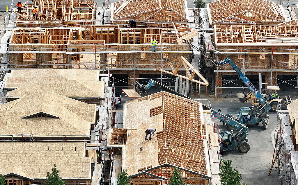 Aerial view of houses under construction with wooden frames, workers on rooftops, and machinery lifting materials at a busy building site.