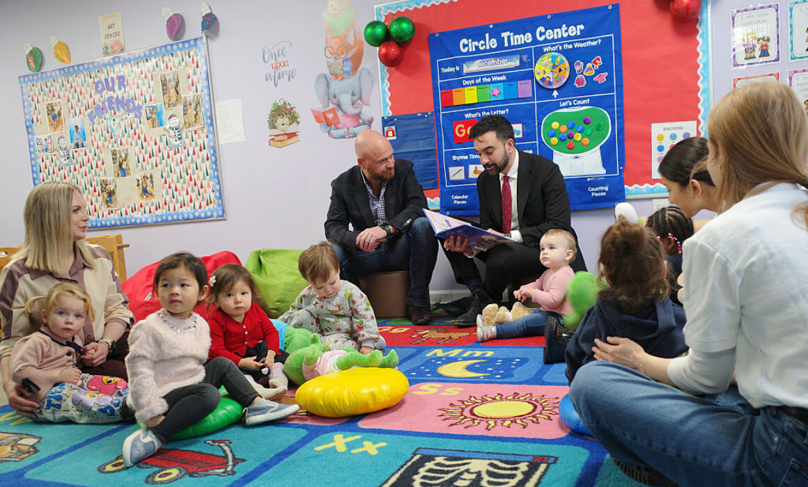 A group of young children and several adults sit in a colorful classroom on a rug, listening to a man in a suit reading from a large book. Classroom posters and decorations are visible on the walls.
