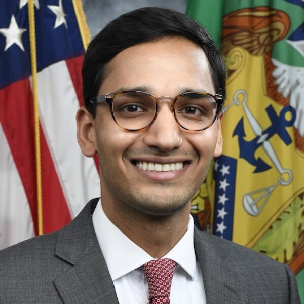 A man wearing glasses, a gray suit, white shirt, and red patterned tie smiles in front of U.S. and government flags.