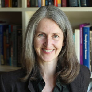 A woman with long gray hair and a dark blazer smiles in front of a bookshelf filled with colorful books.