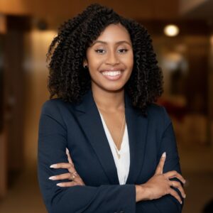 A woman with curly hair wearing a navy blazer and white blouse stands indoors with her arms crossed, smiling confidently at the camera. The background is softly blurred, suggesting an office environment.
