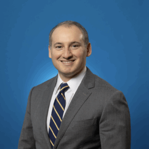 A man in a gray suit, white shirt, and striped tie smiles at the camera against a solid blue background.