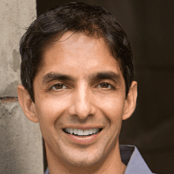 A man with short dark hair, wearing a light gray collared shirt, is smiling at the camera. He is standing next to a rough, concrete wall with a dark background behind him.