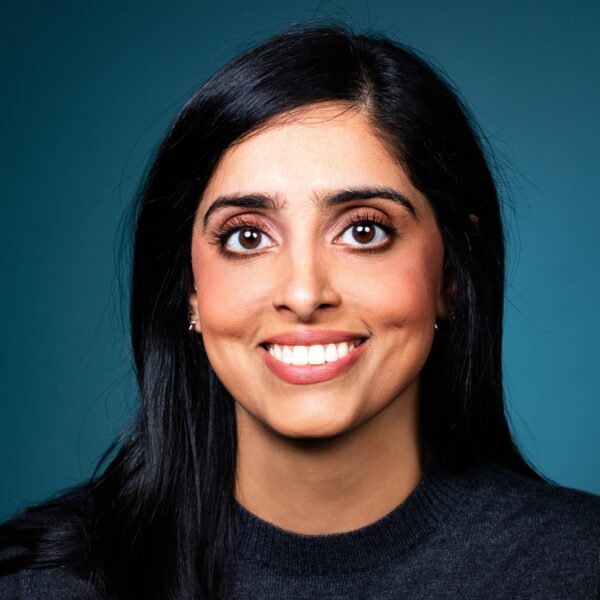 A person with long black hair and brown eyes smiles at the camera. They are wearing a dark sweater, and the background is a solid teal color.