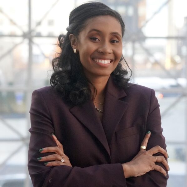 A smiling woman with long dark hair wears a dark blazer and gold jewelry, standing with arms crossed in front of a large window with a geometric frame.
