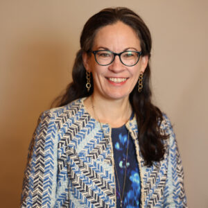 A woman with long dark hair and glasses smiles at the camera. She wears a blue patterned jacket over a dark blue top and gold earrings, standing with her hands resting on a chair against a neutral background.